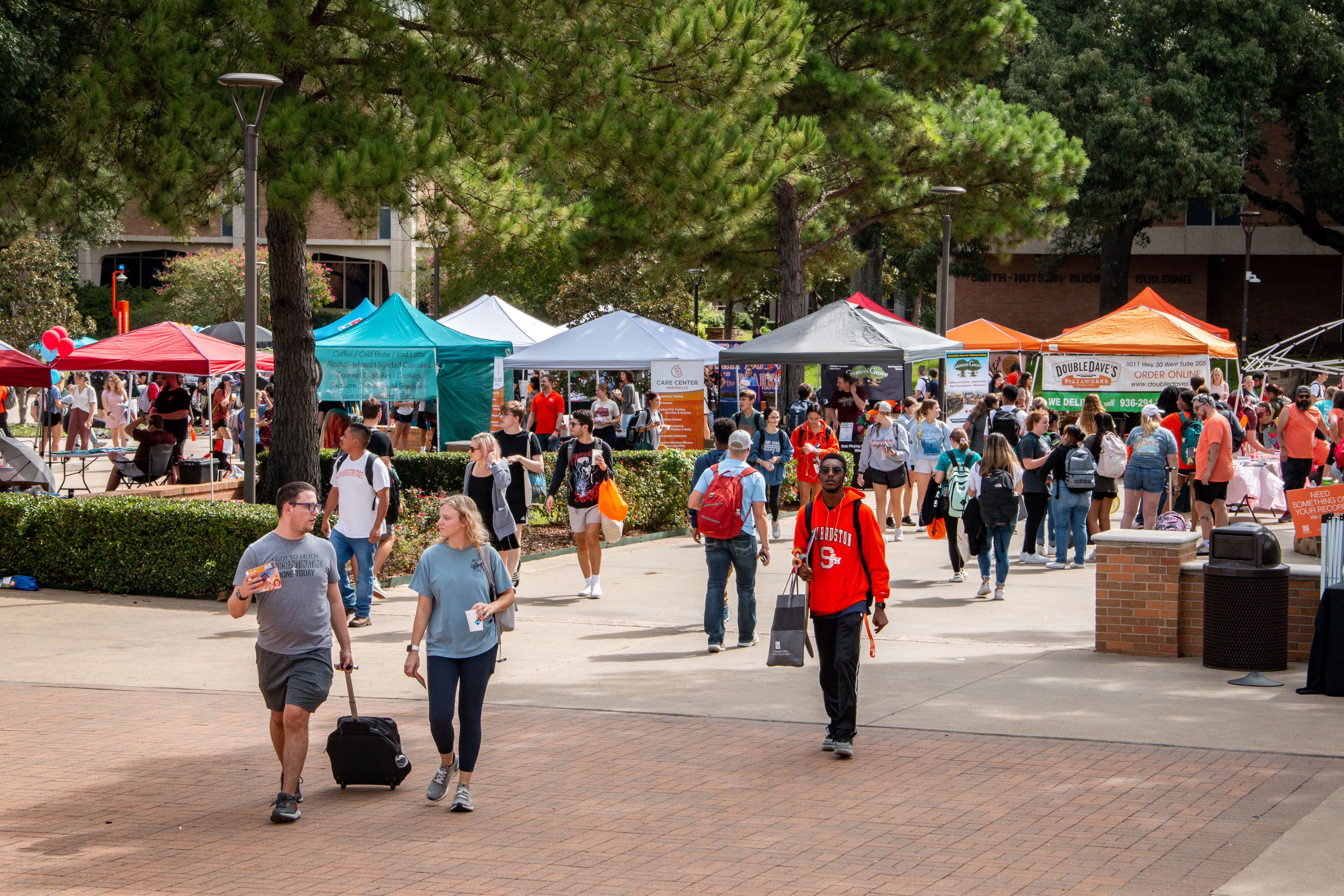 People walking around vendor booths in a tree-dotted plaza.